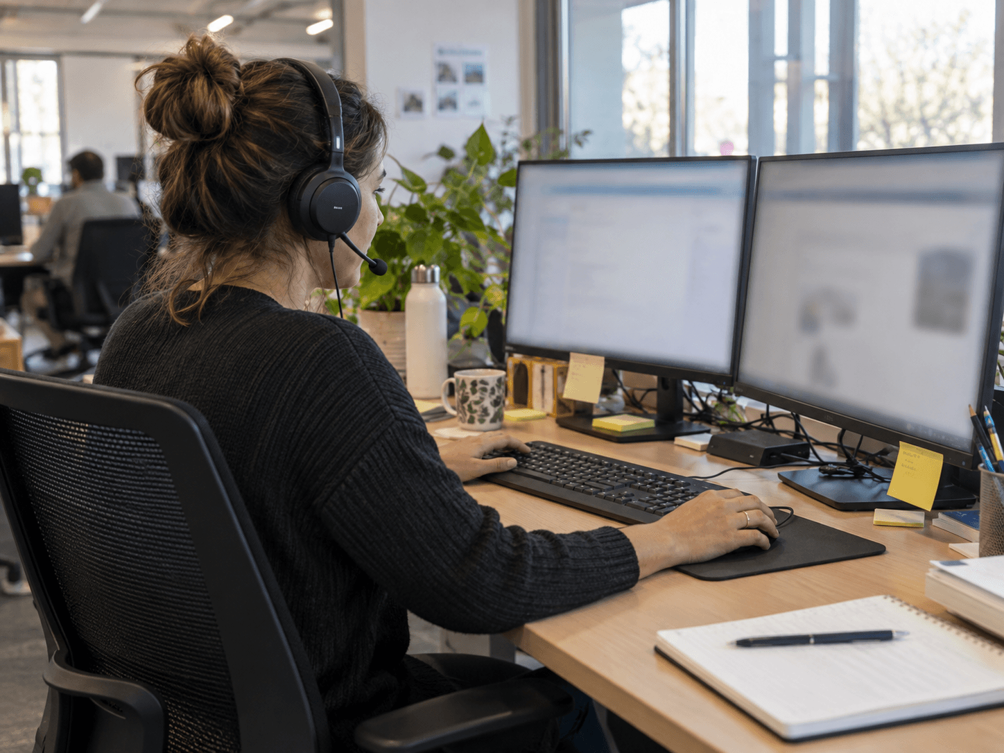 Someone wearing a headset works at two monitors in an office, hands on the keyboard and mouse.