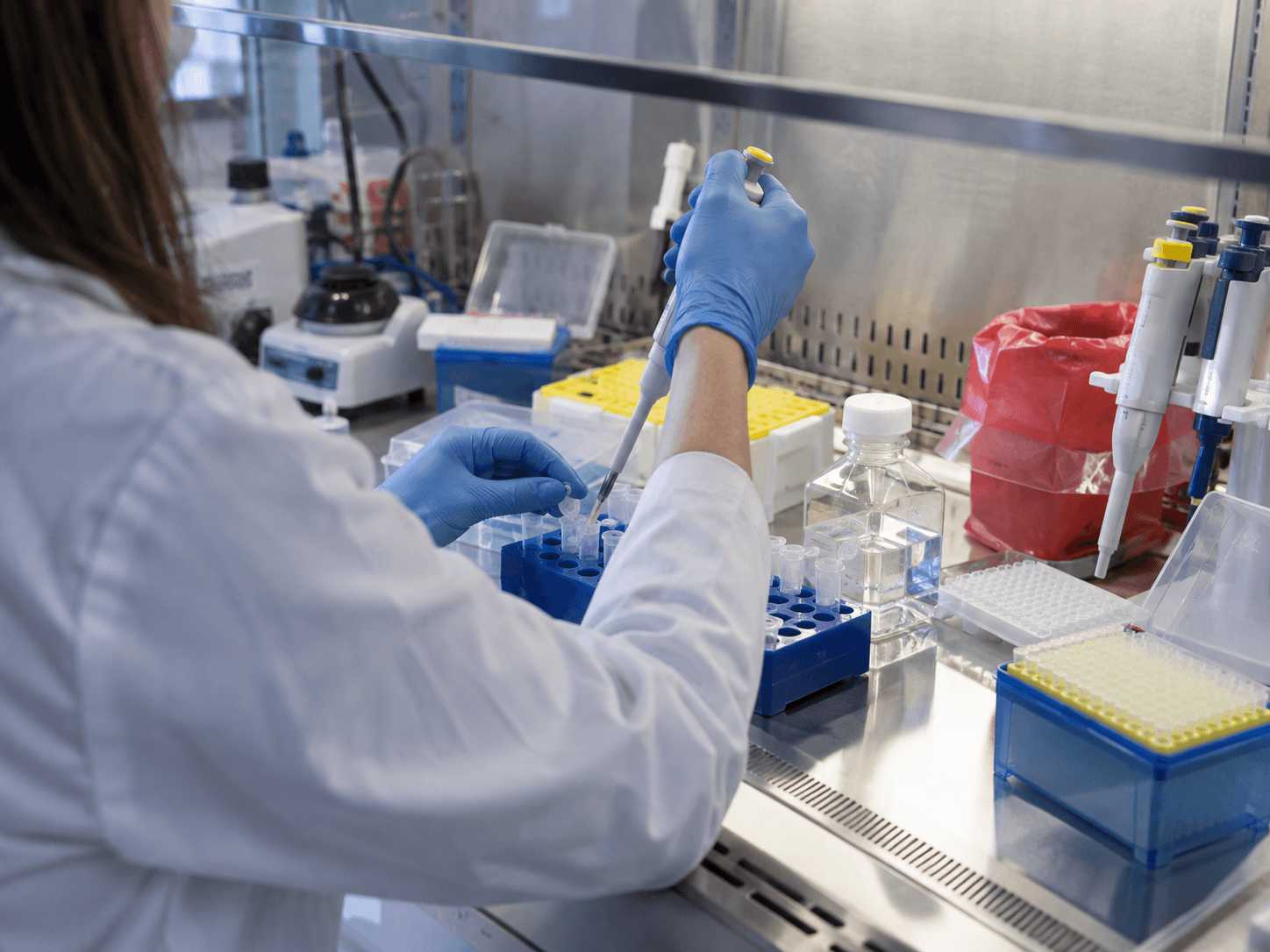 Une technicienne en blouse et gants pipette un échantillon dans un portoir de tubes, sous une hotte de laboratoire.